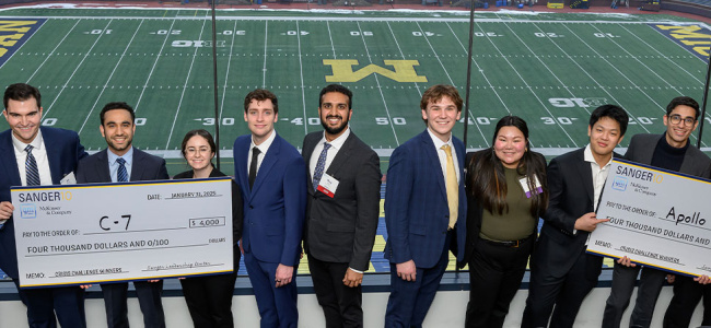 A group of 15 professionally dressed individuals pose with large ceremonial checks in front of an empty Michigan football stadium, exuding pride and accomplishment.