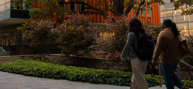 Two people walk along a campus path lined with trees and modern buildings.