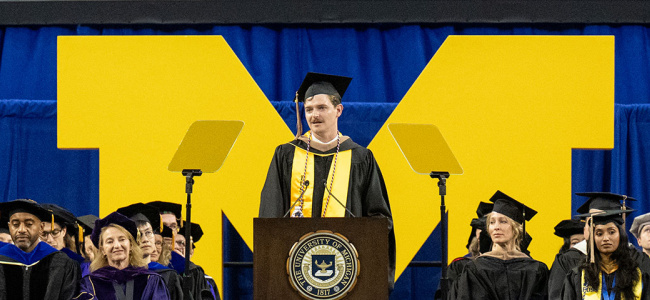 A commencement speaker addresses graduates on stage in front of a large yellow University of Michigan "M".