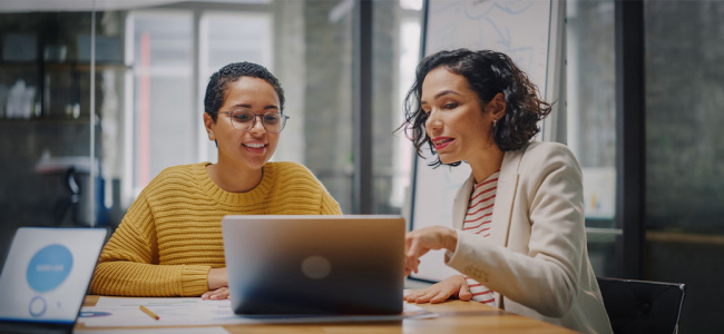 Two colleagues discuss work while looking at a laptop in a modern office.