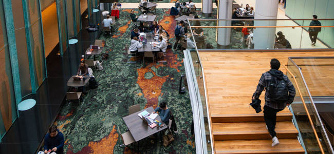 Students studying and socializing in a modern campus common area with tables and stairs.
