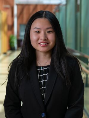 Person in a black blazer smiling in a brightly lit hallway.