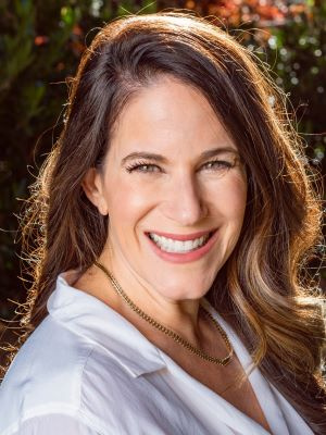 Smiling person with long brown hair wearing a white shirt and gold necklace outdoors.