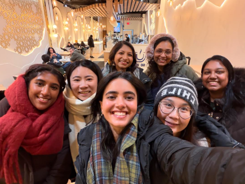 Seven people smiling for a group selfie inside a warmly lit modern café.