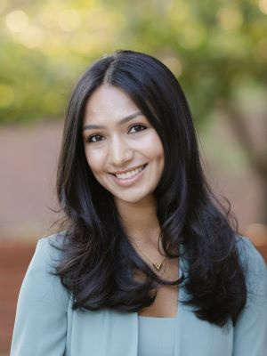 Person smiling outdoors in professional attire with blurred greenery in the background.