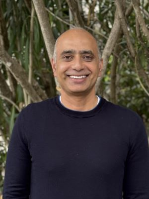 A person in a dark sweater smiles while standing outside in front of trees.