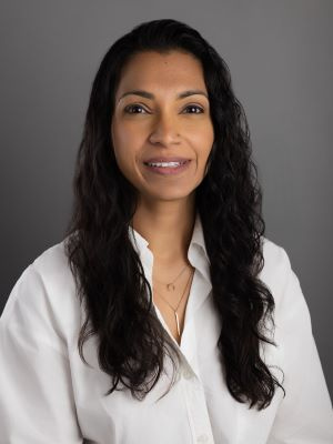 Smiling woman with long dark hair wearing a white blouse against a gray background.