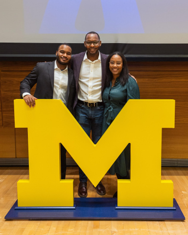 Three people smile behind a large yellow University of Michigan block M logo.