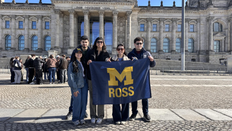 Five people stand in front of a historic building holding a flag reading "M ROSS."