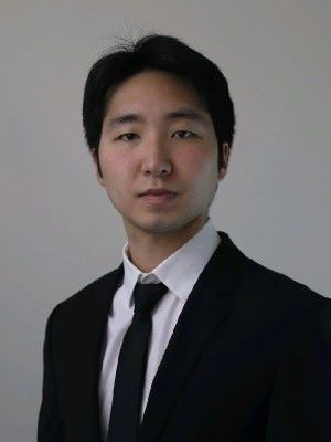 A person wearing a black suit and tie against a plain background.