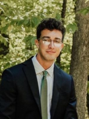 Man in a dark suit and green tie standing outdoors in a wooded area.