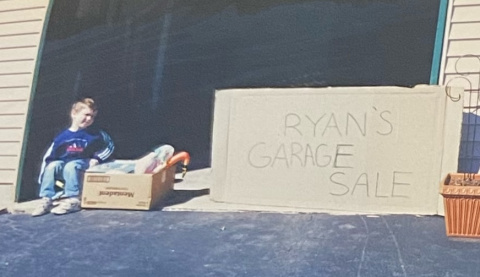 Young child sits by a box of items at a garage sale with a sign reading “RYAN’S GARAGE SALE.”