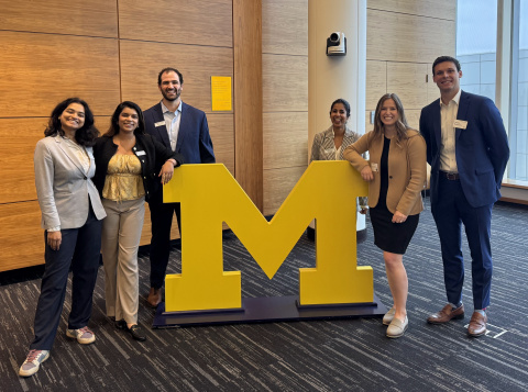 Six people stand smiling around a large yellow University of Michigan block "M" sign.