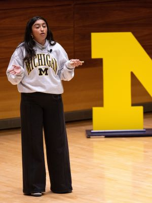 Student wearing a Michigan sweatshirt speaks on stage beside a large yellow block M.