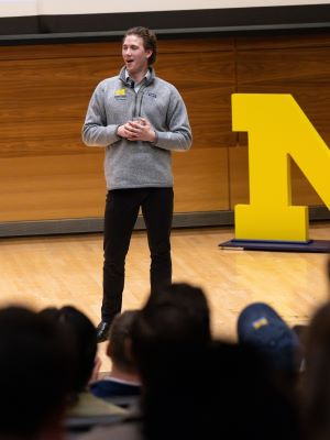 Speaker addressing an audience in an auditorium with a large yellow letter M behind them.