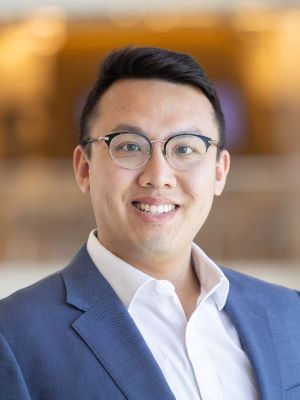 Professional headshot of a smiling person wearing glasses and a navy blazer with a white shirt.