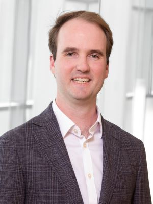 Professional headshot of a man in a checkered blazer and white shirt, smiling.