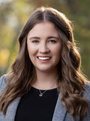 Professional headshot of a smiling woman with long brown hair wearing a black top and grey blazer.