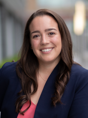 Professional headshot of a woman with long dark hair wearing a navy blazer and pink top.