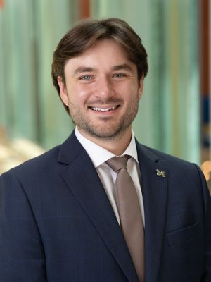 Professional headshot of a smiling man in a dark suit and tie with a lapel pin.