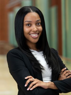 Professional headshot of a smiling woman with long dark hair wearing a black blazer, arms crossed.