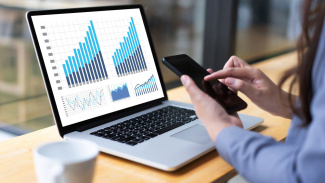 Person checks smartphone while reviewing bar and line graphs on a laptop.