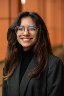 Woman with long dark hair smiling and wearing glasses and a black suit with a black turtleneck sweater