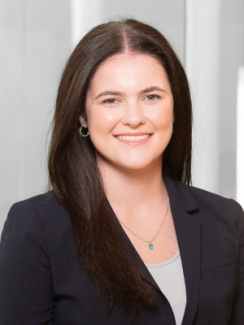 Person with long brown hair smiling, wearing a dark blazer and light top, in an office setting.