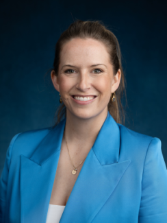 Smiling woman wearing a bright blue blazer against a dark blue background.