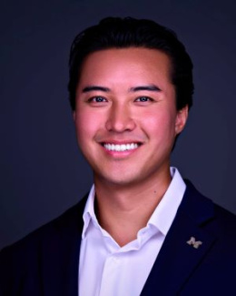 A man smiles, wearing a black blazer and white shirt