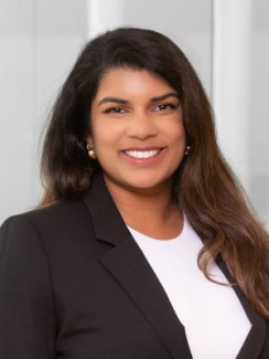 Smiling woman in a black blazer and white top standing in front of a blurred background.