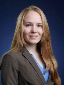 Person in a brown blazer and blue shirt smiling against a dark blue background.