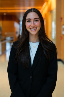 Smiling person with long dark hair wearing a black blazer and white top in an office setting.