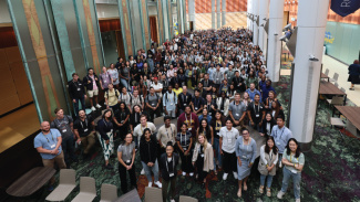 A large crowd of MBA students stand in the Ross Winter Garden beside the large pillars, looking up at the photographer, who is taking the photo from the mezzanine