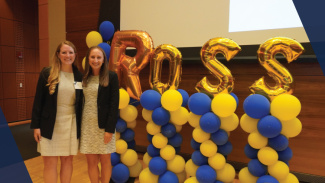 Two women stand side by side with giant balloons that spell "Ross" on a stage