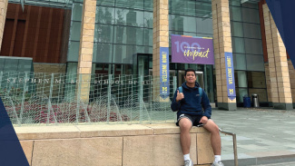 A student sitting on the Ross sign in front of the exterior of the building, smiling and doing a thumbs up gesture