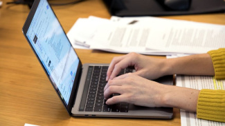Photo of hands typing on a laptop computer. The person is wearing a yellow sweater and you can't see their face. 