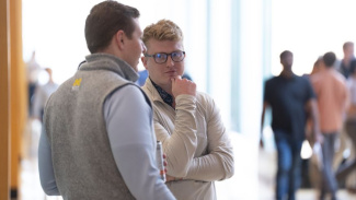 Photo of two male students standing and talking outside of class. One has blond hair and glasses and the other has dark hair and has his back to the camera. Other students are in the background but you cannot see their faces. 