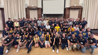 Photo of all of the class members of the newest Michigan Ross Executive MBA class. They are sitting in a large room, many are wearing maize and blue shirts. 