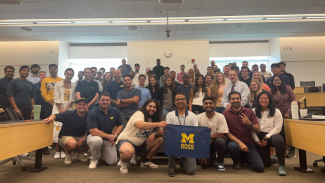 Photo of Weekend MBA students at Michigan Ross all standing together in a classroom holding a maize and blue flag. 