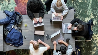 Photo of five Michigan Ross students sitting in the Winter Garden at a table with their computers out. The shot is from up above so you can only see the top of their heads. Two are wearing black sweatshirts, two are wearing gray sweatshirts, and one is wearing a cream colored shirt. 
