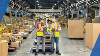 Two people standing in an Amazon warehouse, holding up the Ross flag while wearing safety equipment like reflective vests and hard hats. In the background, there are many boxes.