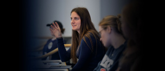 A woman with long brown hair gestures while speaking in a classroom, surrounded by other students. The atmosphere is focused and engaged.