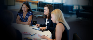 Three women sit at a round table in a well-lit office, engaged in discussion. One woman takes notes on a tablet, creating a focused and professional tone.