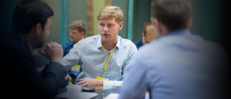 A group of people engaged in a business meeting. The focus is on a person in a light blue shirt with a yellow lanyard, speaking seriously.
