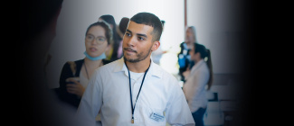 A person in a white shirt with a lanyard stands in focus at an event, pensively listening. Blurred background of people engaged in conversations. Casual atmosphere.