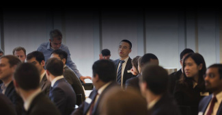 A man in a suit speaks and gestures during a business meeting in a well-lit room. Attendees, mostly in suits, listen attentively, creating a professional atmosphere.