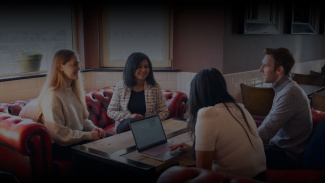 A diverse group of four people engage in a lively discussion around a wooden table in a cozy cafe, with laptops and notebooks, exuding collaboration.