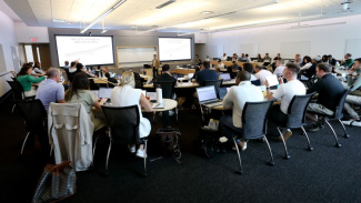Photo of a classroom at Michigan Ross. There are a lot of students sitting and listening to the professor. There are two large screens in the background with the course content displayed. We only see the backs of the students. 