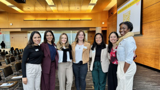 Photo of seven female Weekend MBA students standing in a room with their arms around each other. They are looking at a camera and smiling. 
